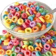 Bowl of colorful fruity cereal rings with spoon and scattered pieces on white surface